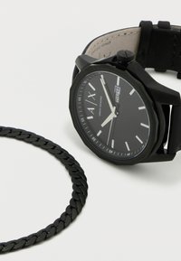 Črna analogna ura s teksturiranim okroglim številčnico, belimi številčnicami ure in usklajenim pletenim zapestnico. Ilustrira zaslon datuma in oktagonalno luneto.