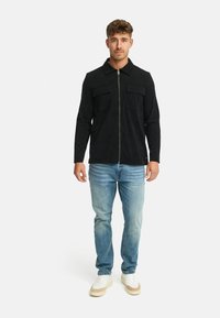 Sort zip-up jakke med to brystlommer, ensartet tekstur, parret med lyseblå jeans og hvide sneakers med beige detaljer.