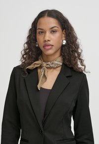 Černý blazer s klopami, vrstvený přes tmavý top bez rukávů. Modelka má na sobě světle hnědý šátek s vzorovaným akcentem a stříbrné náušnice.