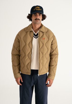 Brown quilted jacket with diamond pattern, side pockets, and a front zipper. Worn over a cream knit sweater and dark trousers; cap with logo.