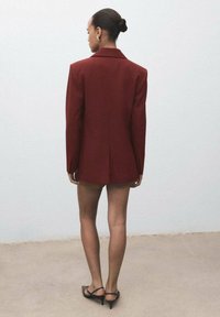 Bordeaux oversized blazer met gestructureerde schouders, enkele split aan de achterkant en gladde stof, gedragen over shorts. Model staat met de rug naar de camera.