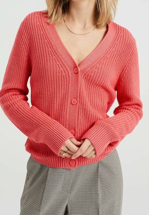 Femme portant un cardigan corail rose en tricot boutonné à manches longues et un pantalon gris à carreaux, mains jointes devant.