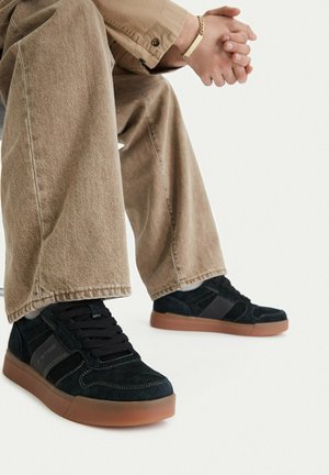 Persona sentada con las manos entrelazadas, vistiendo pantalones beige y zapatillas negras con suelas marrones sobre un fondo blanco.