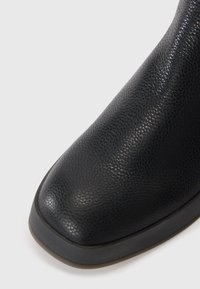 Botín negro de cuero hasta el tobillo con un acabado texturizado, punta redonda y suela de goma baja. Presenta un borde suave en el cuello.