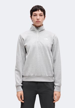Osoba ubrana w jasnoszary pulower z suwakiem do połowy oraz logo North Face i czarne spodnie, stojąca frontem na jednolitym tle.