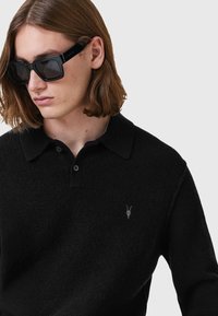 Mustapaita, jossa on kauluskanta ja kaksi nappia. Rakenteellinen kangas. Rinnassa pieni brodeerattu emblem.