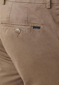 Chinos marrones con cierre de botón, bolsillo con detalle de costura y una etiqueta de marca que dice "Hackett". Tejido suave con un ligero brillo.