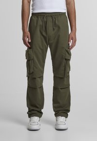 Pantalones cargo verde oliva con cintura elástica, múltiples bolsillos laterales y un corte ajustado. Llevados con zapatillas deportivas blancas.