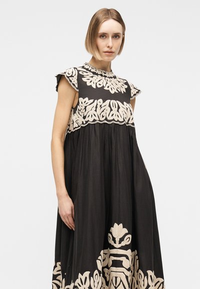Γυναίκα με κοντά μαλλιά που φοράει μαύρο αμάνικο φόρεμα με κεντήματα σε εκρού χρώμα στο μπούστο και το τελείωμα, στέκεται μπροστά σε απλό φόντο.