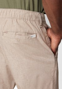 Light beige pants made of textured fabric, featuring an elastic waistband, back pocket, and a small tag. Focus on practicality and comfort.