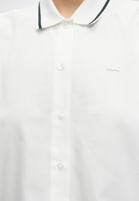 Chemise blanche à col avec un liseré noir sur le col, devant boutonné, et un petit logo crocodile blanc brodé sur la poitrine.