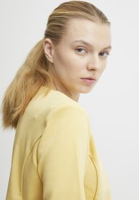 Žlutý dlouhý rukáv s pevným střihem, s jemným vycpáváním na ramenou a hladkým, texturovaným materiálem; modelka má světlé vlasy.
