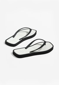 Zwarte slipper met dunne bandjes, voorzien van een gestructureerde witte zool met golvenpatronen en een subtiele logo. Rechthoekige vorm met een platte basis.