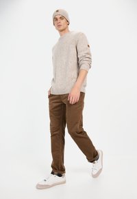 Crème geklede trui met geribbelde manchetten en kraag, bruine corduroy broek en witte sneakers met beige accenten; model draagt een gebreide muts.