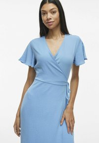 Vaaleansininen kääntömekko, joka on valmistettu tekstuurimateriaalista. Mekossa on lyhyet liehuvat hihat ja vyötäröllä solmittava nauha, joka mahdollistaa säädettävän istuvuuden.