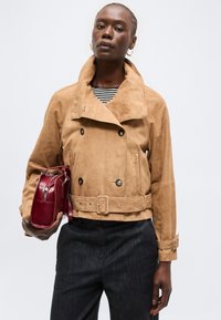 Γυναίκα που φοράει διπλό παλτό από σουέντ ταμπά με ζώνη, μαύρο παντελόνι, ριγέ μπλουζάκι, χρυσά κρίκους σκουλαρίκια, κρατά κόκκινη δερμάτινη τσάντα.