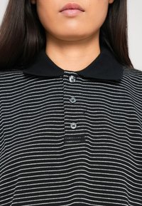 Femme portant un polo rayé noir et blanc avec trois boutons et un col noir uni, montrée des épaules aux lèvres.