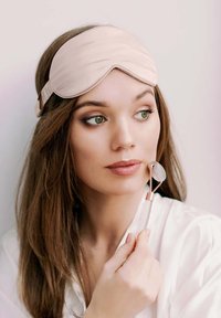 Hellrosa Satin-Schlafmaske, die auf langen braunen Haaren getragen wird; hält einen roségoldenen und klaren Gesichtsroller; weiche weiße Kleidung vor neutralem Hintergrund.