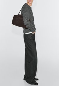 Person in dunklem grauem Pullover und weitem schwarzen Hosen, trägt eine große dunkelbraune Schultertasche und steht vor weißem Hintergrund.