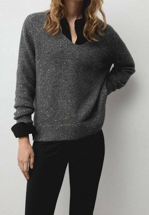 Personne portant un pull gris foncé moucheté par-dessus une chemise noire avec un pantalon noir sur un fond clair uni.