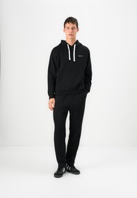 Black sweatshirt with a hood and white drawstrings, featuring a front pocket and logo detail. Paired with matching black pants and sneakers.
