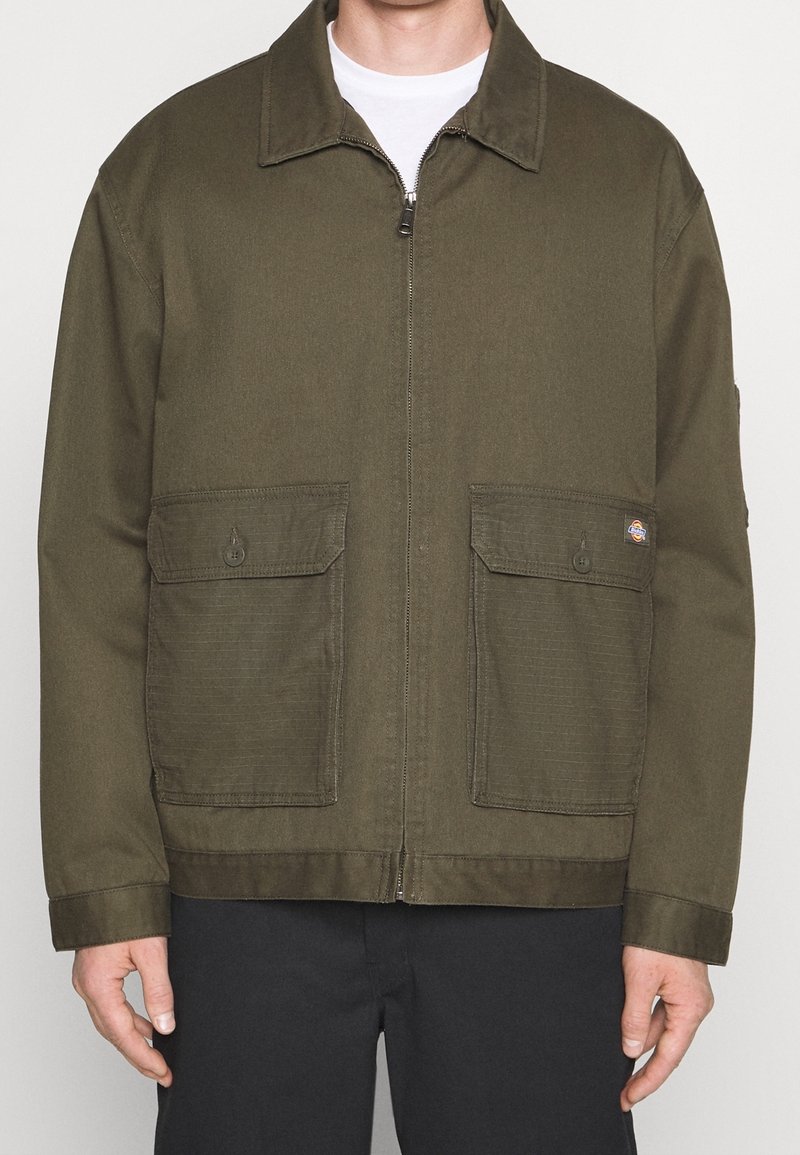 Homme portant une veste zippée vert olive avec deux poches avant boutonnées par-dessus une chemise blanche, pantalon noir visible sous la taille.