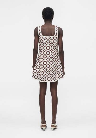 Γυναίκα που φοράει αμάνικο, πλεκτό φόρεμα μέχρι το γόνατο με καφέ, λευκά και γαλάζια τετραγωνισμένα σχέδια, στέκεται με την πλάτη προς το φακό σε απλό φόντο.