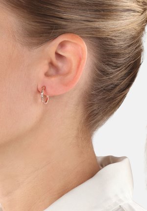 Oreja de mujer con un pequeño pendiente de aro dorado retorcido, tono de piel claro y cabello recogido, vistiendo una camisa blanca con cuello.