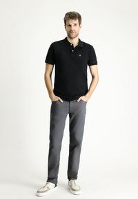 Camiseta polo de algodón negra con cuello, combinada con pantalones grises y zapatillas blancas. Presenta un pequeño logo en el pecho.