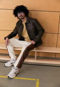 Καφέ δερμάτινο μπουφάν πάνω από μουσταρδί φούτερ, συνδυασμένο με κρεμ φόρμα με καφέ ρίγες και γκρι αθλητικά παπούτσια σε ξύλινο πάγκο.