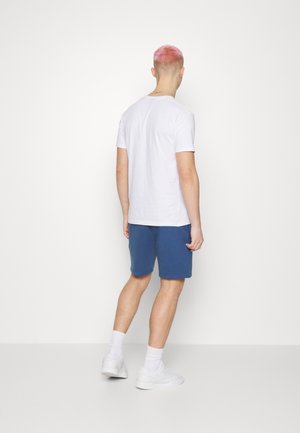 Jeune personne aux cheveux courts roses portant un t-shirt blanc uni, un short bleu, des chaussettes blanches et des baskets blanches, debout, vue de dos.