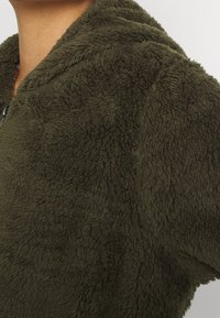 Oliwkowa polarowa kurtka z miękką, teksturowaną powierzchnią, z kapturem i zapięciem na zamek z przodu. Tkanina wygląda na grubą i przytulną.