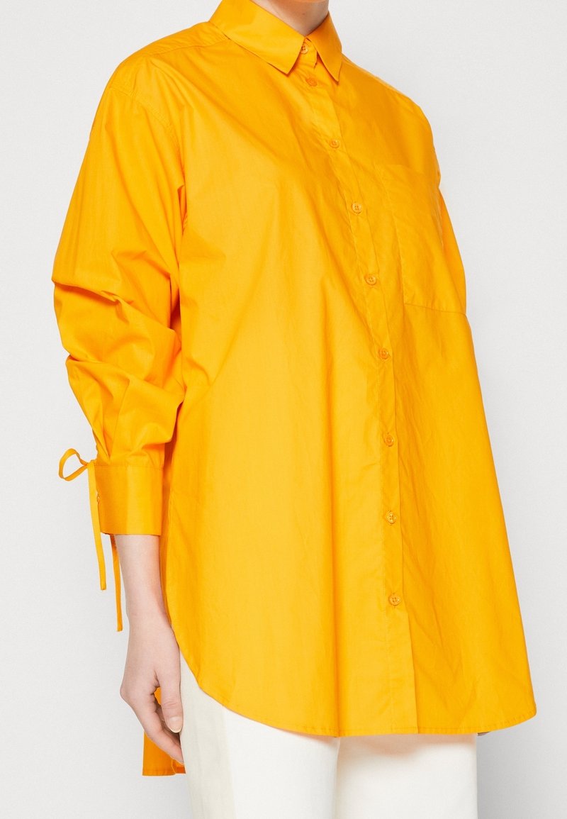 Chemise orange vif à manches longues avec col et boutons, coupe ample, poignets noués, portée avec un pantalon blanc, montrée de la moitié du torse vers le bas.