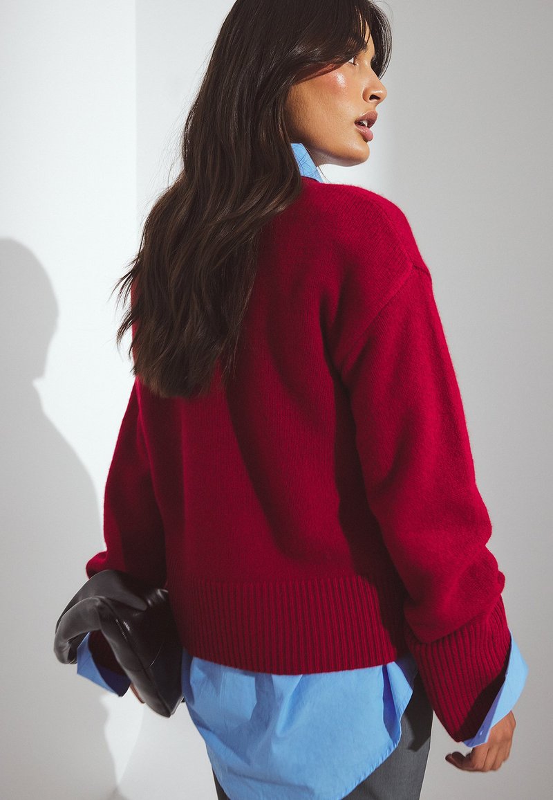 Raudonas megztinis su raukšlėtu apvadu ir plačiomis rankovėmis, uždėtas ant šviesiai mėlynos marškinėlių. Laiko juodą odinį rankinį krepšį. Lygus paviršius.