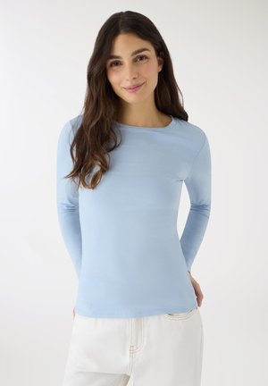 Kobieta z długimi ciemnymi włosami, ubrana w jasnoniebieską bluzkę z długim rękawem i białe spodnie, stoi na tle jednolitego białego tła, delikatnie uśmiechając się.