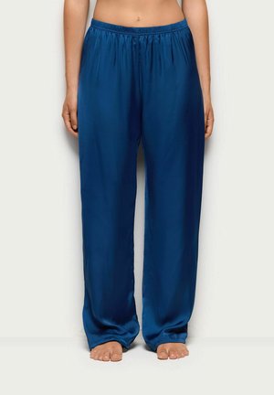 Persona que lleva puestos unos pantalones de satén azul oscuro, sueltos y brillantes, de pie descalza sobre un fondo claro y sencillo.