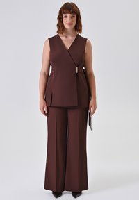 Donna che indossa un top avvolgente senza maniche di color marrone scuro con cintura e pantaloni larghi coordinati, in piedi davanti a uno sfondo chiaro e semplice.