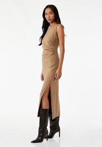 Bodysculpting beige maxi-kjole med sideskæring, tætsiddende design, rund halsudskæring og en glat tekstur, parret med knæhøje sorte støvler.