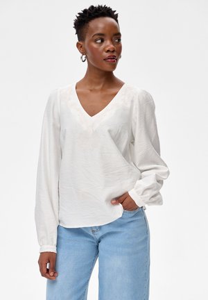 Vrouw met kort haar die hoepeloorbellen draagt, een witte blouse met lange mouwen en V-hals, en lichtblauwe spijkerbroek, terwijl ze opzij kijkt met één hand in haar broekzak.