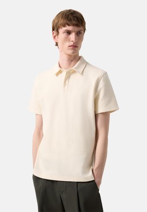 Jonge man met kort bruin haar draagt een crème korte mouw polo shirt en donkere broek, staat met handen in de zakken tegen een witte achtergrond.