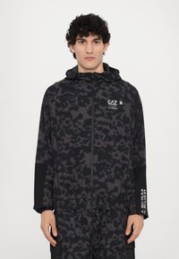 Black athletic jacket with a textured, abstract pattern. Features a hood, zip closure, and logo on the chest. Slim fit with long sleeves.