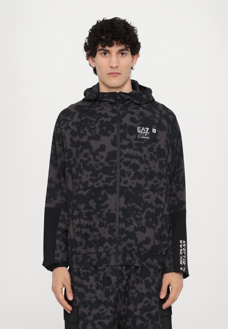 Svartidrettsjakke med en teksturert, abstrakt mønster. Har hette, glidelås og logo på brystet. Slim fit med lange ermer.
