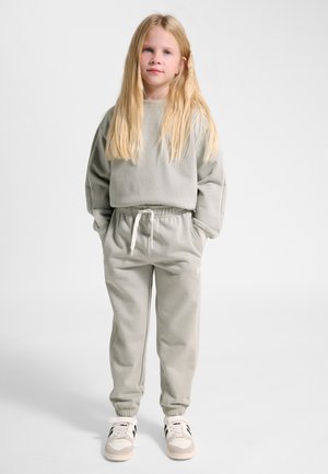 Junges Mädchen mit langen blonden Haaren steht mit Händen in den Taschen, trägt hellgraue Sweatpants, passenden Sweatshirt und weiße Sneaker vor weißem Hintergrund.