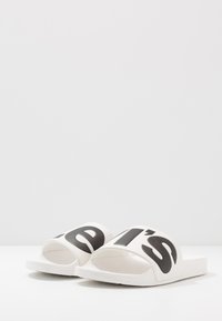Sandalias de playa blancas con letras negras gruesas a lo largo de la tira ancha, suela plana y área del dedo del pie curvada. Material suave con diseño minimalista.