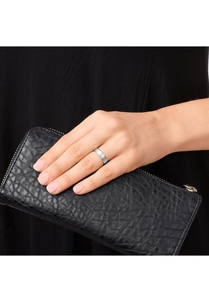 Silberner Ring mit zwei horizontalen Rillen, gehalten in einer Hand, die auf einer strukturierten schwarzen Lederbörse mit Reißverschluss ruht.