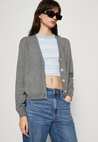 Siv cardigan iz mehkega materiala, z gumbi spredaj in rebrastimi manšetami, v kombinaciji z modrim prugastim crop topom in hlačami z visokim pasom.