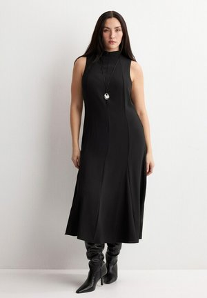 Femme aux longs cheveux noirs portant une robe midi noire sans manches, des bottines à talons pointues noires, et un collier pendentif, debout contre un mur blanc.