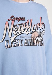 Ljusblå sweatshirt med fet grafisk tryck som visar texten "League New York" och "Classic Athletics", samt en illustration av en sko.