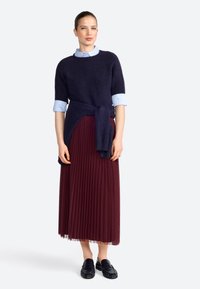 Navy knitted sweater over a light blue collared shirt, paired with a burgundy pleated skirt and black loafers. Soft texture and layered design.