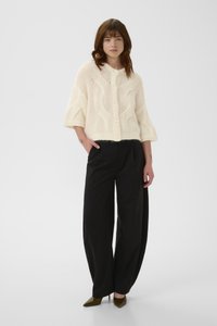 Λευκό πλεκτό ζακέτα με υφή ανάγλυφου σχεδίου cable, κοντά μανίκια και κουμπιά μπροστά, συνδυασμένο με φαρδιά μαύρη παντελόνα και μυτερές ψηλοτάκουνες.